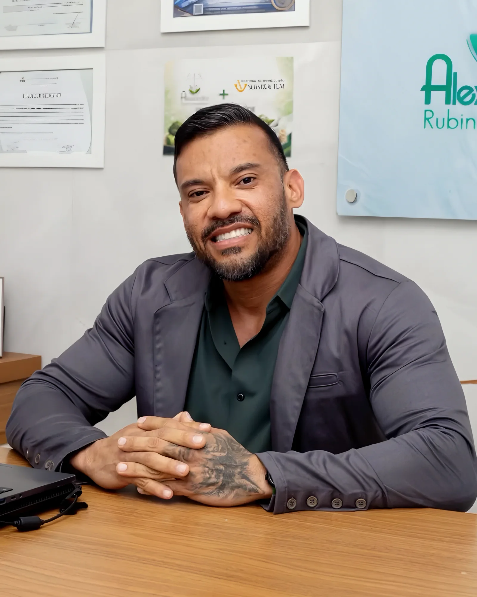Dr. Alexsandro Rubin sorrindo, sentado à mesa do consultório com as mãos cruzadas, de blazer cinza e camisa verde, com certificados e o painel do logotipo ao fundo