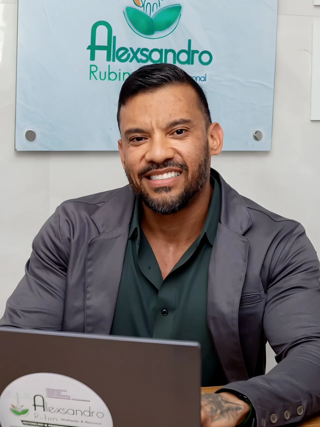 Dr. Alexsandro Rubin sorrindo à mesa do consultório, com o notebook aberto, de blazer cinza e camisa verde, diante do painel de vidro com o logotipo