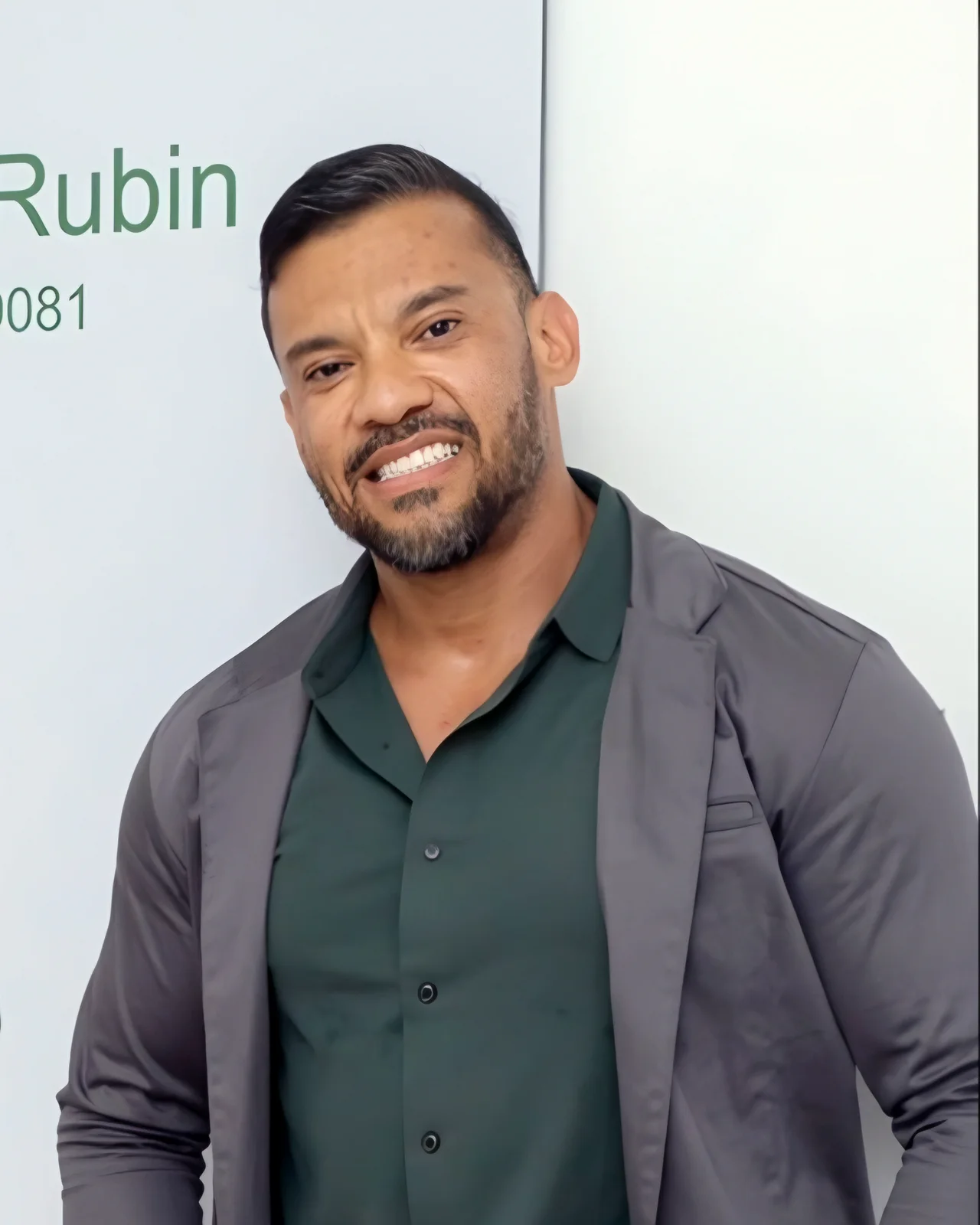 Dr. Alexsandro Rubin de pé, sorrindo, junto à porta do consultório com a placa do nome, de blazer cinza e camisa verde