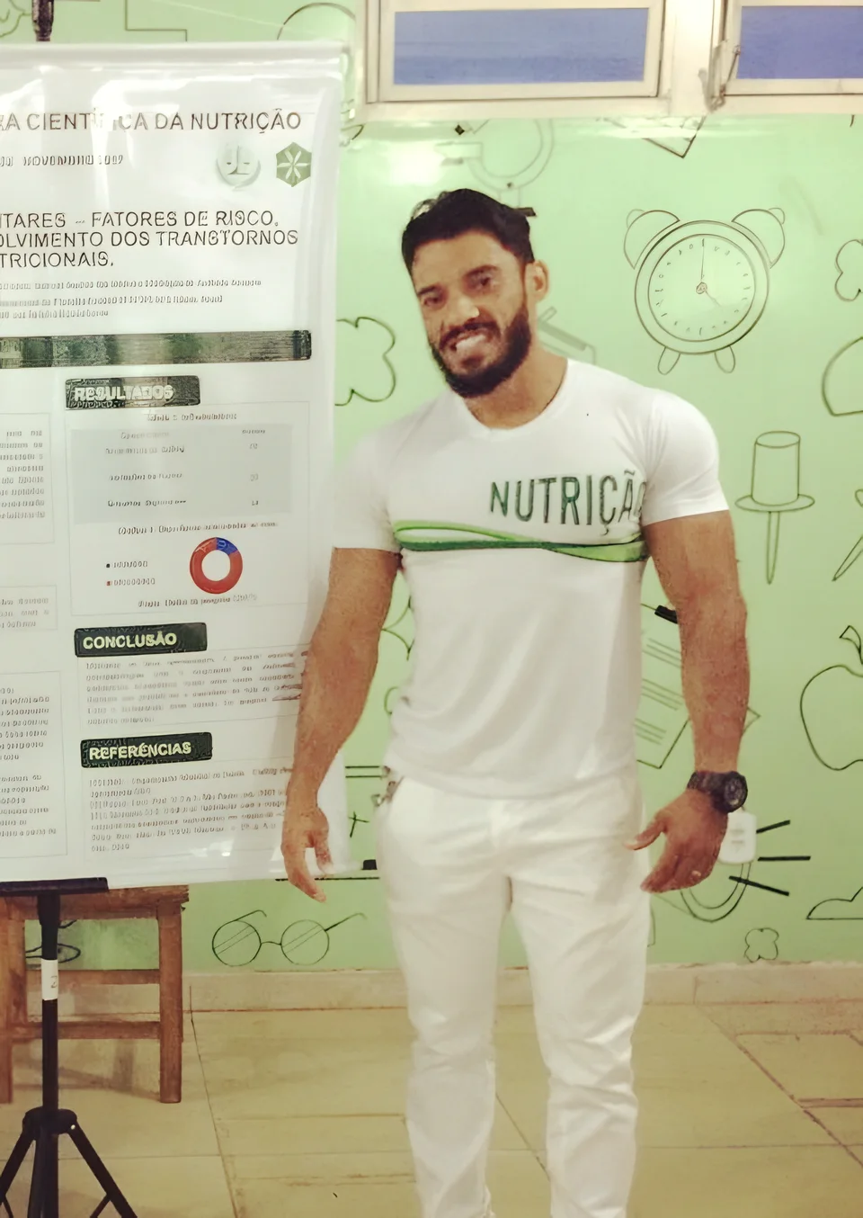 Alexsandro Rubin com camiseta de Nutrição ao lado do banner de um artigo científico apresentado na faculdade