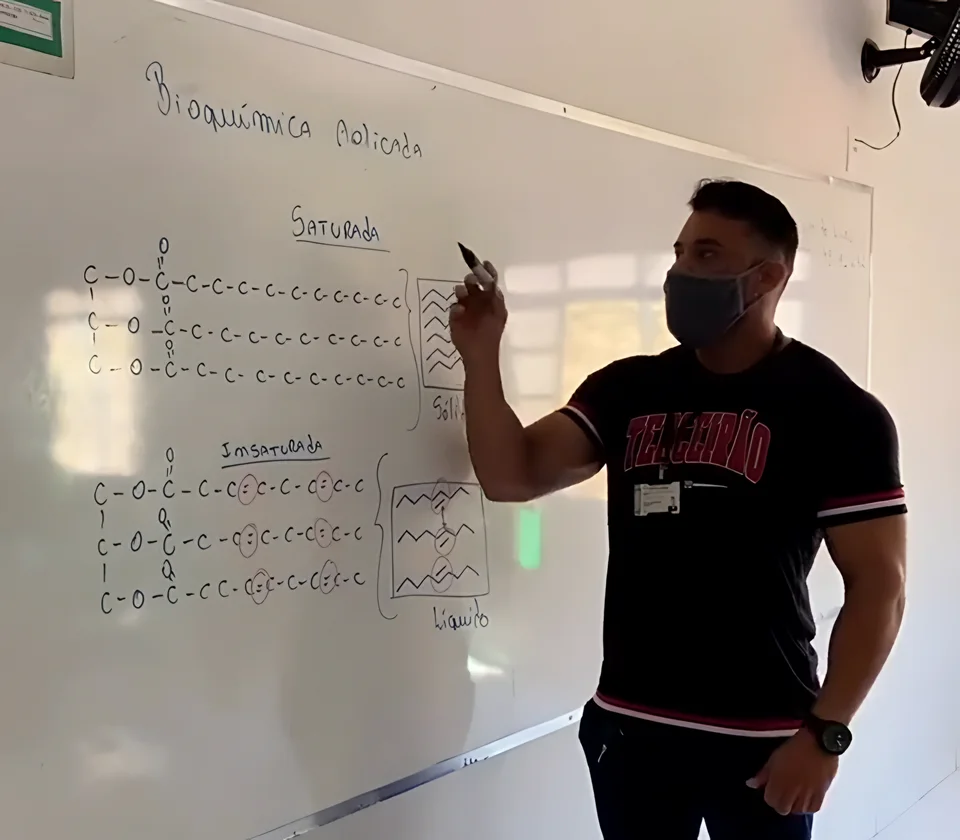 Alexsandro Rubin de máscara escrevendo fórmulas de bioquímica no quadro branco durante uma aula